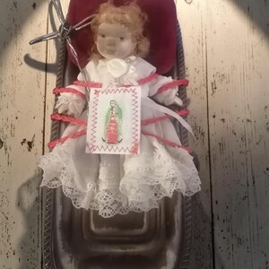 May include: A porcelain doll with curly hair wearing a white lace dress, secured with pink cord to a dark metal tray with a red velvet cushion. A small rectangular tag with a printed image of the Virgin of Guadalupe is attached to the front.
