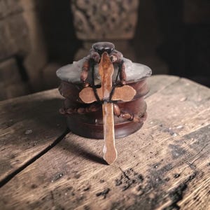 May include: A wooden cross with a dark cord hangs in front of a small, round, wooden container. The container has a dark, round lid and is encircled by a string of wooden beads. The cross is light brown and has a simple design.