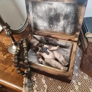 May include: A wooden box with a distressed finish houses a small doll adorned with a black cross. A rosary with a detailed cross lies beside the box. The box's interior features aged paper with printed text, creating a vintage aesthetic.