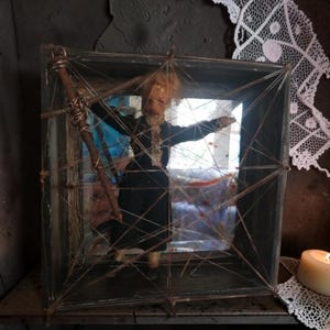 May include: A doll with light brown hair wearing a black outfit is positioned inside a square wooden frame. The frame is crisscrossed with thin, light-colored string, creating a web-like pattern across the front. A dark, textured stick is attached to the left side of the frame, and a piece of white lace fabric hangs from the upper right corner.
