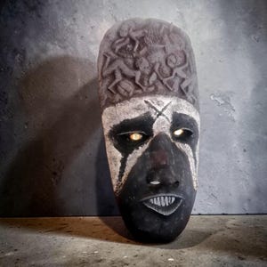 Op de afbeelding: Een gesneden houten masker met een donkere, getextureerde afwerking. Het masker heeft een zwart gezicht met witte accenten rond de ogen en het voorhoofd, en een gesneden ontwerp aan de bovenkant. De ogen gloeien geel. De mond is open en toont tanden.