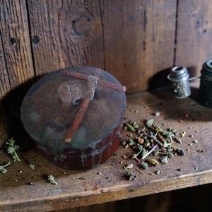 May include: A round, dark-coloured jar with a translucent, wax-like lid featuring embedded cinnamon sticks and a dusting of brown powder. Beside the jar lies a small pile of dried green herbs and botanical pieces, with a metal tea infuser nearby.