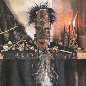 Op de afbeelding: Een gesneden houten masker met een gedetailleerd gezicht, versierd met veren, kralen en edelstenen. Het masker heeft een donkerbruine afwerking met ingewikkelde gravures en wordt tentoongesteld op een houten plank met zwarte kant en gedroogde planten. Twee zwarte kaarsen en een brandende kaars zijn ook aanwezig.