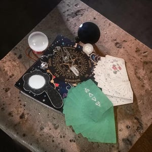 May include: A collection of divination tools including a crystal ball, a black orb, a Ouija board, a deck of tarot cards, a crystal pendant, and green cards. The Ouija board has the words "YES", "NO", and "REPHRASE" printed on it.