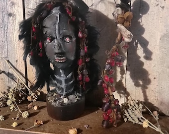 Puede incluir: Un busto esculpido de tonos oscuros con un diseño de pecho esquelético, plumas negras y detalles de cuentas rojas. Junto al busto se encuentra un bastón alto y delgado adornado con racimos de cuentas rojas y moradas, pequeñas campanas y elementos decorativos similares a huesos.