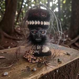 Puede incluir: Una escultura artesanal de rostro oscuro con ojos naranja brillantes, adornada con un collar de cuentas y un tocado decorativo. La escultura tiene una textura negra similar a la piel alrededor del cuello y se exhibe sobre una base de madera.