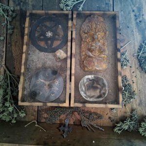 Op de afbeelding: Een houten doos met zand, een zwarte pentagramschijf met een kleine ster, een bruin reliëf van een figuur onder een boom, een ronde metalen plaat met een stierenschedelontwerp, een kleine steen en een metalen kom, met een kruishanger eronder.