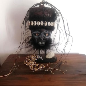 May include: A black mask with a stylized face, decorated with white shells and dark fur. The mask is mounted on a stand, with a beaded necklace and a small stone on a wooden surface. Long, thin strands extend from the mask.