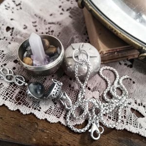 Puede incluir: Un collar plateado con un colgante redondo que se abre para revelar una piedra oscura. El collar está junto a un pequeño recipiente redondo plateado lleno de cristales y piedras, y un recipiente plateado en forma de manzana.