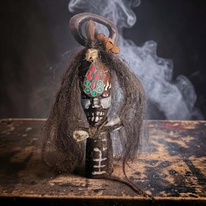 Peut inclure: Poupée en bois artisanale avec un visage sculpté, de longs cheveux foncés et un pendentif décoratif avec des accents rouges et turquoise. La poupée a un aspect unique et vieilli, avec une base marron foncé et des détails complexes.