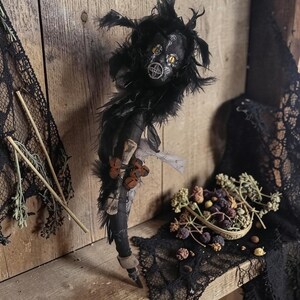 May include: A handcrafted doll featuring a black fabric body adorned with dark feathers, a round head with yellow eyes, and white painted markings. The doll is decorated with a cluster of small, rusted metal bells and a light-coloured fabric wrap.