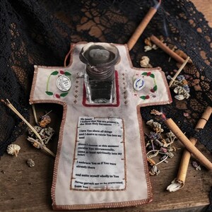 May include: A cross-shaped religious item with a small ink bottle, text, and decorative elements. The cross is made of fabric with embroidered details and small metal medallions. Incense sticks and dried flowers surround the cross.