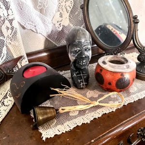 Op de afbeelding: Een verzameling decoratieve items, waaronder een zwarte houten kandelaar met een rode kaars, een kleine messing bel, een gebeeldhouwde stenen buste en een rood-oranje decoratieve kom. Op de achtergrond een kleine spiegel.