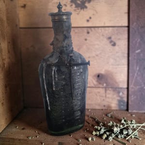 May include: A dark, translucent glass bottle with embossed lettering reading "PHILLIPS MILK OF MAGNESIA". The bottle neck is coated in hardened white wax, featuring a metal cap, a thin chain, and a long, dark, rusted iron nail attached to the front.