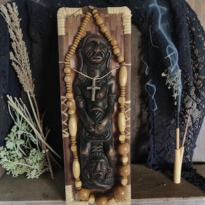 Op de afbeelding: Een donkere houten sculptuur van een figuur met een kruisvormige hanger, omlijst door geweven bamboe. Een kralenketting met houten kralen en langwerpige kralen omringt het beeldhouwwerk. Wierookrook stijgt op in de achtergrond.