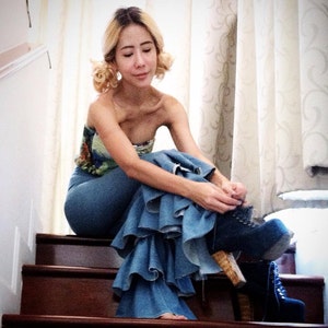 May include: A woman wearing a blue denim skirt with multiple layers of ruffles. The skirt is a flared style and the woman is sitting on a wooden staircase. She is wearing a floral print top and has blonde hair.