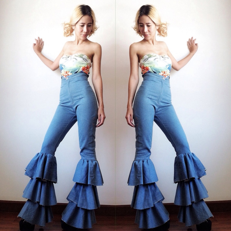 May include: A woman is wearing a blue denim high-waisted pants with a multi-colored floral print top. The pants have a wide leg and multiple layers of ruffles at the bottom.