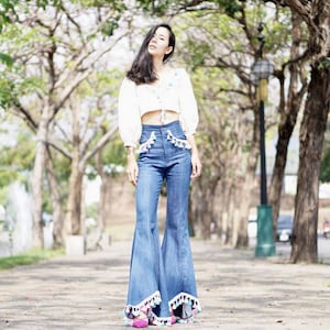 May include: A woman in a white crop top with floral embroidery and flared blue jeans. The jeans have tassel detailing on the pockets and hem. The background shows a tree-lined path, typical of a park or garden.