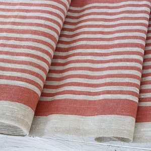 May include: Folded fabric with horizontal stripes in a muted red-orange and off-white. The fabric appears to be linen or a similar natural fibre, with a slightly textured surface. The edges are finished with a neutral border.