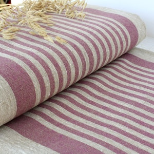 May include: A folded piece of fabric featuring a striped pattern in deep plum and natural beige. The textile has a visible woven texture and is decorated with a small cluster of dried oat stalks resting on the top surface of the folded material.