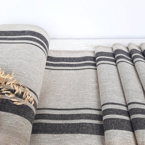 May include: A roll of natural beige fabric featuring a woven texture and prominent dark charcoal grey stripes running horizontally. The fabric is partially unrolled, displaying a flat section and several folded layers, with a small sprig of dried oats.