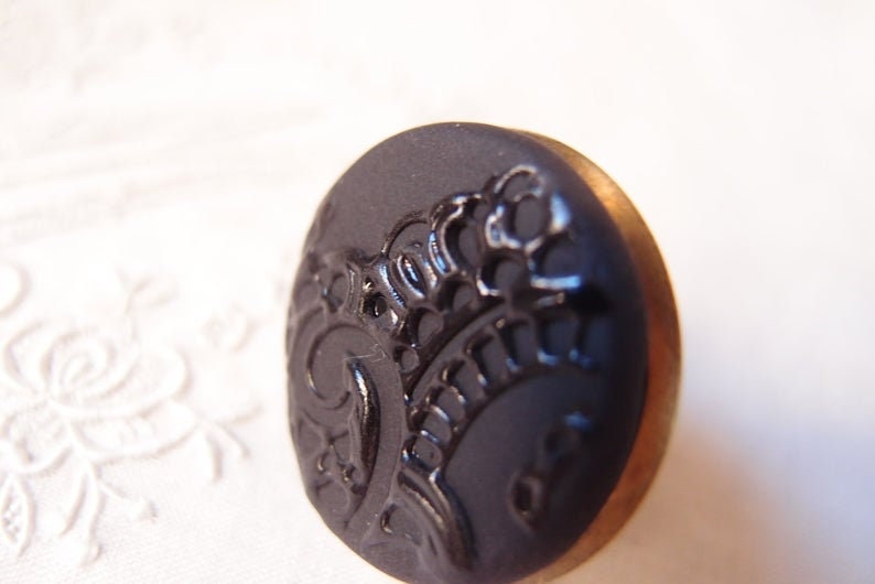 Pu&ograve; includere: Un bottone nero con un bordo dorato e un design decorativo. Il design &egrave; un motivo floreale stilizzato.