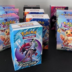 May include: Multiple Pokémon trading card game boxes are displayed. The boxes are primarily blue, with vibrant graphics of Pokémon characters and the Pokémon logo. The boxes are labeled "Prismatic Evolutions" and "Journey Together".