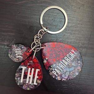 May include: A keychain with three pendants. The pendants are red and black with glitter and white text that reads "FUCK", "THE", and "PATRIARCHY". The keychain has a silver key ring and chain.