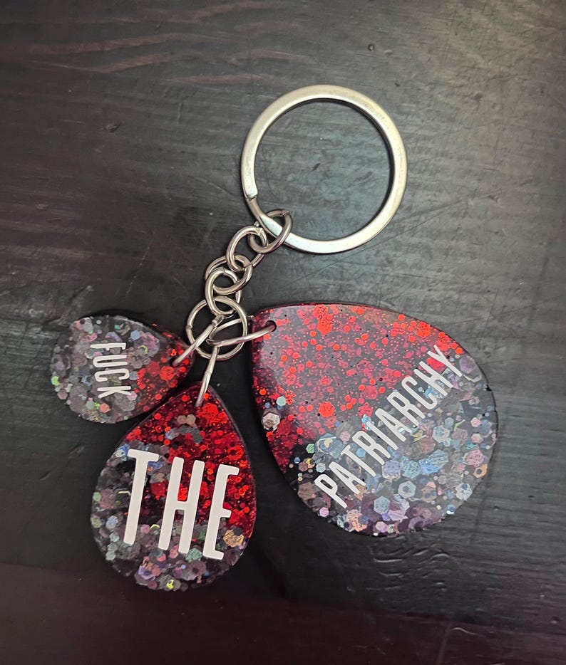 May include: A keychain with three pendants. The pendants are red and black with glitter and white text that reads "FUCK", "THE", and "PATRIARCHY". The keychain has a silver key ring and chain.