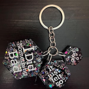 May include: A keychain with three black, glitter-filled, hexagonal charms. The charms have white text that reads "Bad Something I Did", "it just felt so", and "good". The keychain has a silver ring and chain.
