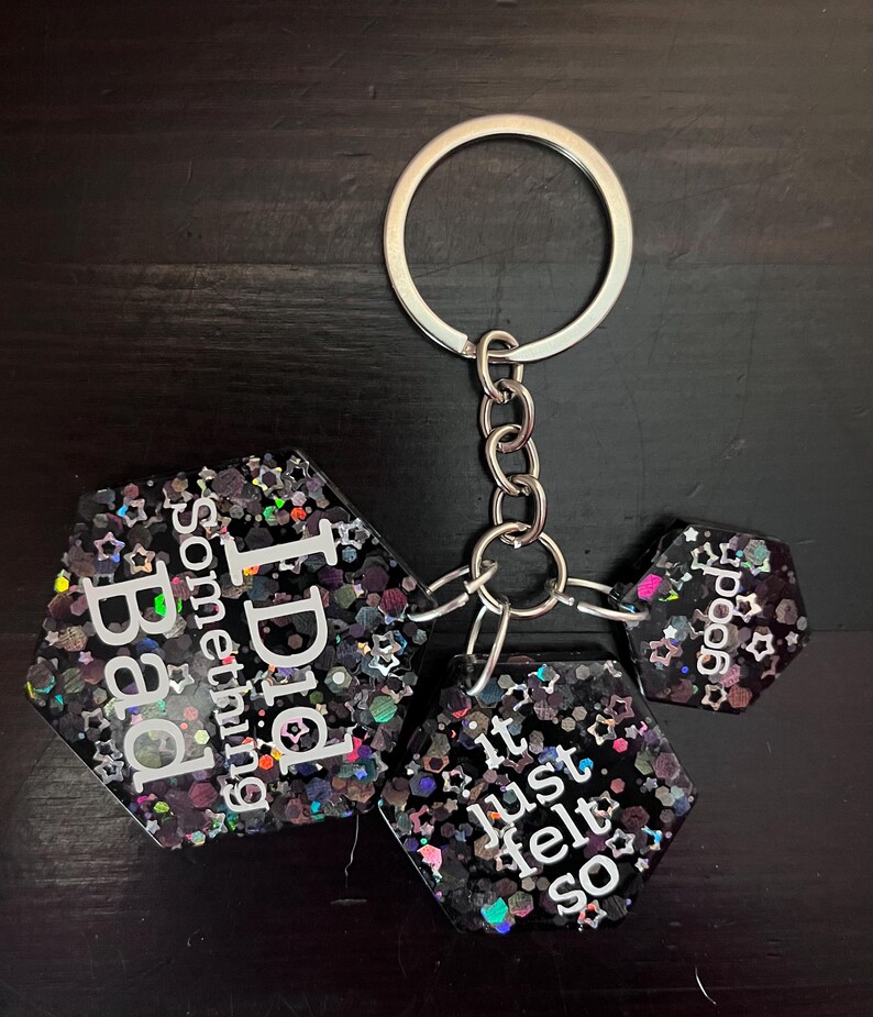 May include: A keychain with three black, glitter-filled, hexagonal charms. The charms have white text that reads "Bad Something I Did", "it just felt so", and "good". The keychain has a silver ring and chain.