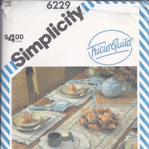 May include: Simplicity pattern 6229 by Tricia Guild. The image shows a table setting with a floral patterned table runner, placemats, plates, napkins, and a teapot. The color scheme includes blue, gold, and beige.