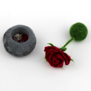 Puede incluir: Un cuenco de fieltro gris lleno de cuentas de colores y una rosa de fieltro rojo con un tallo y un capullo de fieltro verde.