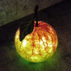 Puede incluir: Una manzana de fieltro roja y amarilla que brilla, con una hoja verde. La manzana está iluminada desde adentro, creando un brillo cálido.