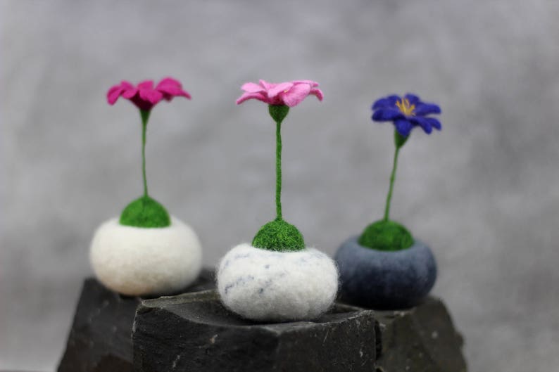 Puede incluir: Tres arreglos florales de fieltro en macetas blancas, grises y blancas. Las flores son rosas, azules y moradas.