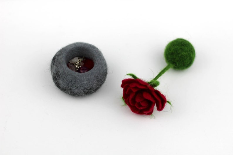 Puede incluir: Un cuenco de fieltro gris lleno de cuentas de colores y una rosa de fieltro rojo con un tallo y un capullo de fieltro verde.