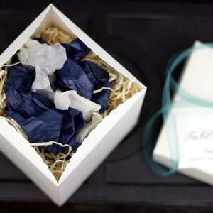 Puede incluir: Una caja de regalo blanca abierta con un borde festoneado, llena de papel de seda azul y blanco. La caja est&aacute; sobre una superficie negra. El papel de seda est&aacute; arrugado y tiene un aspecto natural y org&aacute;nico.