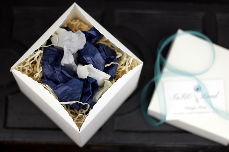 Puede incluir: Una caja de regalo blanca abierta con un borde festoneado, llena de papel de seda azul y blanco. La caja est&aacute; sobre una superficie negra. El papel de seda est&aacute; arrugado y tiene un aspecto natural y org&aacute;nico.