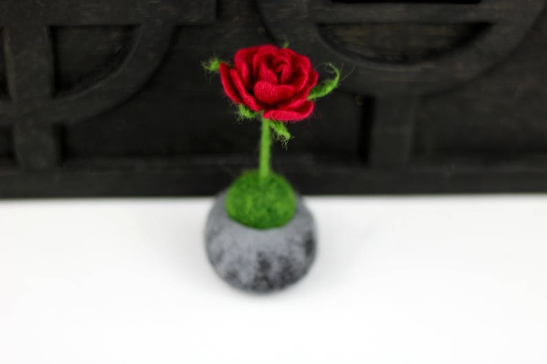 Puede incluir: Una sola rosa roja de fieltro con hojas y tallo de fieltro verde, en una peque&ntilde;a maceta de fieltro gris con musgo de fieltro verde.
