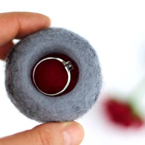 Puede incluir: Una caja de anillo de fieltro gris con un interior de fieltro rojo. La caja de anillo est&aacute; siendo sostenida por una mano.
