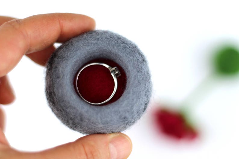 Puede incluir: Una caja de anillo de fieltro gris con un interior de fieltro rojo. La caja de anillo est&aacute; siendo sostenida por una mano.