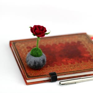 Puede incluir: Una rosa roja de fieltro con hojas verdes de fieltro en una maceta de fieltro gris se encuentra sobre un diario de cuero rojo cerrado con un bol&iacute;grafo plateado al lado.