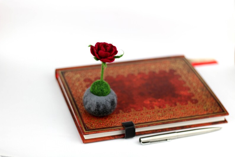 Puede incluir: Una rosa roja de fieltro con hojas verdes de fieltro en una maceta de fieltro gris se encuentra sobre un diario de cuero rojo cerrado con un bol&iacute;grafo plateado al lado.