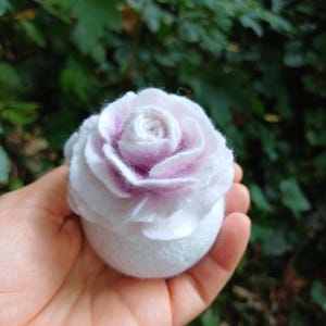 Puede incluir: Una rosa de fieltro blanca y morada. La rosa de fieltro tiene una base redonda y pétalos en capas, con un tono morado más oscuro en el centro. La rosa se sostiene en una mano sobre un fondo verde borroso.