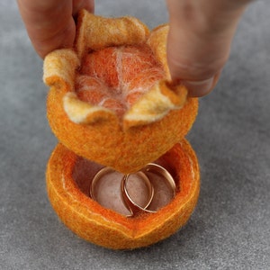 以下が含まれることがあります： フェルトで作られたオレンジ型のリングボックス。蓋を持ち上げると、中に2つのゴールドリングが現れます。ボックスは鮮やかなオレンジ色で、表面はざらざらしています。内側は明るいオレンジ色で、リングはゴールドです。