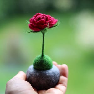Puede incluir: Una rosa de fieltro con tallo y hojas verdes, sentada sobre una base de piedra gris.
