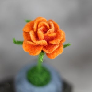 Puede incluir: Una sola rosa de fieltro naranja con hojas y tallo de fieltro verde. La rosa est&aacute; sentada sobre una base de fieltro gris.
