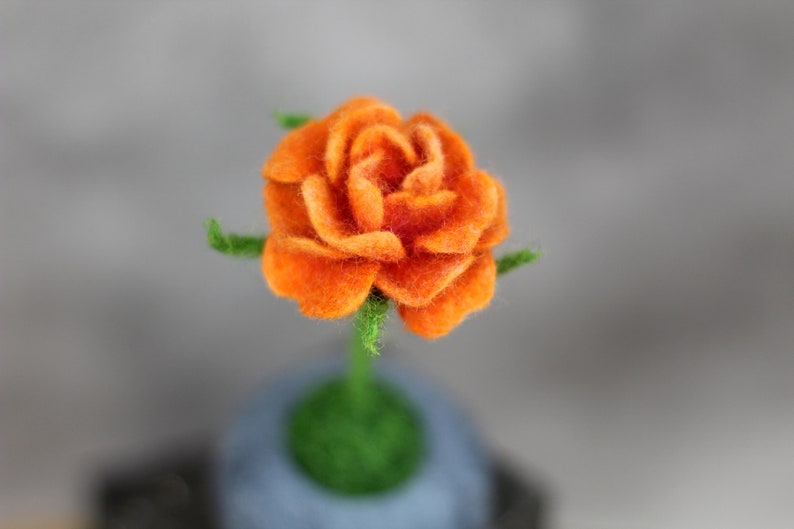 Puede incluir: Una sola rosa de fieltro naranja con hojas y tallo de fieltro verde. La rosa est&aacute; sentada sobre una base de fieltro gris.