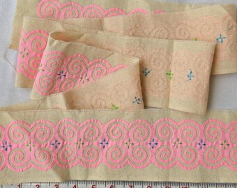 May include: Several strips of beige fabric with a repeating pink spiral and cross design. A ruler is placed along the bottom edge, showing measurements in inches. The fabric has a hand-stitched appearance.