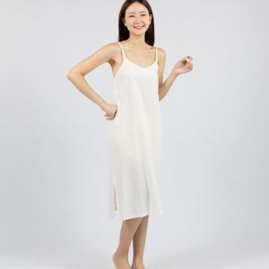 May include: A woman wearing a white sleeveless midi dress with a relaxed fit. She is smiling and standing on a white background.
